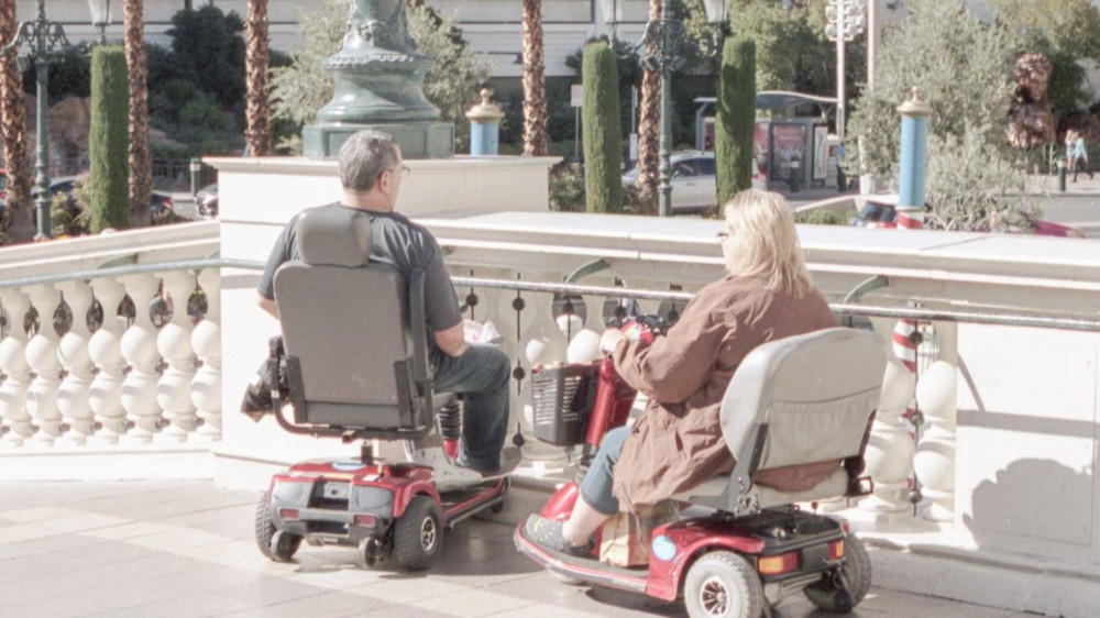 Nainen ja mies istuvat sähköpyörätuoleissa. He katselevat valkoisen aidan taakse.
